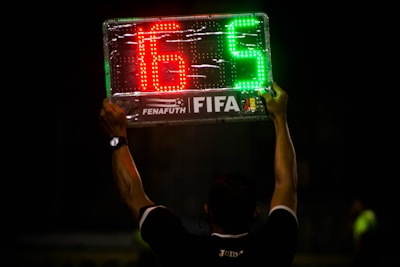 A person is holding up an electronic substitution board displaying the numbers 16 in red and 5 in green, typically used in football matches to indicate player changes.