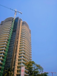 A tall, modern high-rise building is under construction. The structure features rounded balconies and a sleek design. A green construction crane is prominently positioned alongside the building. Trees with green foliage are visible at the base, and there is a clear blue sky as the backdrop.