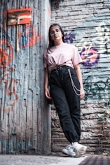 A young person wearing glasses and casual clothing leans against a graffiti-covered urban wall. The scene features a mix of textures and colors from the street art and worn surfaces, creating a vibrant, urban vibe.