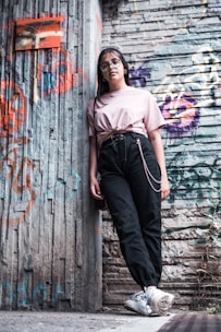 A young person wearing glasses and casual clothing leans against a graffiti-covered urban wall. The scene features a mix of textures and colors from the street art and worn surfaces, creating a vibrant, urban vibe.