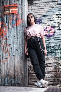 A young person wearing glasses and casual clothing leans against a graffiti-covered urban wall. The scene features a mix of textures and colors from the street art and worn surfaces, creating a vibrant, urban vibe.