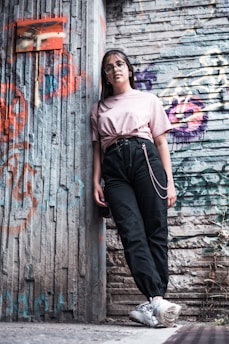 A young person wearing glasses and casual clothing leans against a graffiti-covered urban wall. The scene features a mix of textures and colors from the street art and worn surfaces, creating a vibrant, urban vibe.