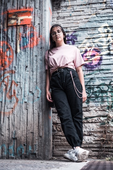 A young person wearing glasses and casual clothing leans against a graffiti-covered urban wall. The scene features a mix of textures and colors from the street art and worn surfaces, creating a vibrant, urban vibe.