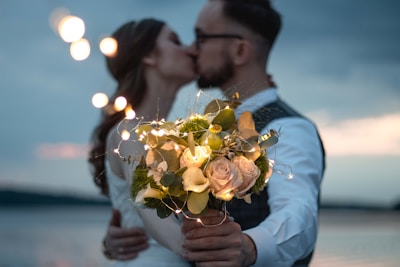 A couple is sharing a romantic kiss near the water, with the focus on a bouquet of flowers they are holding. The bouquet includes pink roses and is decorated with string lights, creating a warm, glowing effect. The background features a serene body of water and a slightly cloudy sky.