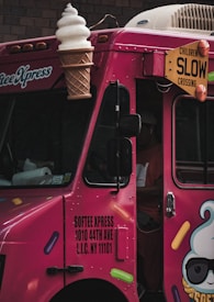 A bright pink ice cream truck with a large ice cream cone decoration on top. The truck has colorful sprinkles illustrations and a sign displaying the text 'SOFTEE XPRESS 1010 44TH AVE L.I.C. NY 11101'. There is a yellow warning sign that says 'CHILDREN SLOW CROSSING'.