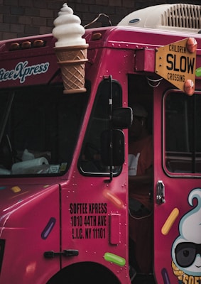 A bright pink ice cream truck with a large ice cream cone decoration on top. The truck has colorful sprinkles illustrations and a sign displaying the text 'SOFTEE XPRESS 1010 44TH AVE L.I.C. NY 11101'. There is a yellow warning sign that says 'CHILDREN SLOW CROSSING'.