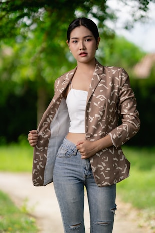 A person is standing outdoors, wearing a patterned brown blazer over a white top and distressed blue jeans. The background consists of lush green trees, creating a vibrant, natural atmosphere.
