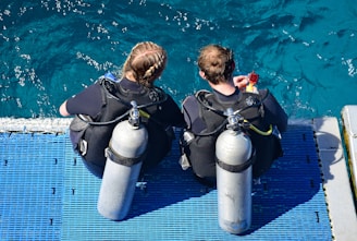 Two people wearing scuba diving gear are seated on a blue platform at the edge of the water. They are equipped with oxygen tanks and wetsuits, preparing to dive into the clear, turquoise sea.