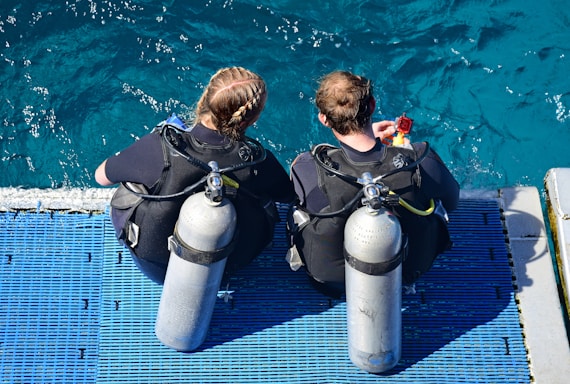 Two people wearing scuba diving gear are seated on a blue platform at the edge of the water. They are equipped with oxygen tanks and wetsuits, preparing to dive into the clear, turquoise sea.