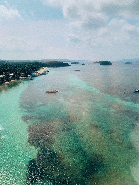 A serene coastal landscape featuring crystal-clear waters in shades of turquoise and green. Several small boats float gently on the water near a lush shoreline lined with dense palm trees. In the distance, a series of small islands rise against the horizon, under a sky dotted with fluffy clouds.