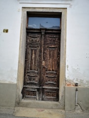 A weathered, wooden double door set in a light-colored wall. The door features intricate carvings and a small, rectangular window above it. Signs of age and wear are visible on the wood and around the door frame, with some peeling paint on the wall.