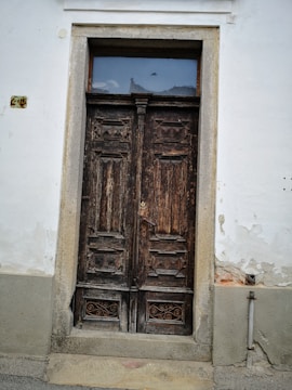 A weathered, wooden double door set in a light-colored wall. The door features intricate carvings and a small, rectangular window above it. Signs of age and wear are visible on the wood and around the door frame, with some peeling paint on the wall.