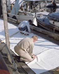Two men are working on a dock near several boats. One man is kneeling and holding a tool while working on a large piece of white material spread out on the wooden dock. Boats and various nautical equipment are visible in the background, suggesting a harbor or marina setting.