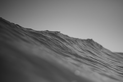 A black and white photograph depicting the ocean with gentle waves under a calm sky. The surface of the water shows texture and movement, with light reflecting off the wave peaks.