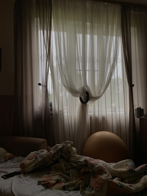 Sheer curtains hang from a window, allowing diffused light to enter the room. A black tie holds the curtains together in the center. A bed with a colorful, patterned duvet appears unmade in the foreground, and a remote control rests on it. To the right of the bed, an exercise ball is positioned near the window, and various small objects are visible on a bedside table.