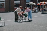 Two people engage in an outdoor activity in a courtyard. One is handing an object to the other in what appears to be a playful setting; a group of children in wheelchairs watch from a distance. The scene is lively with bright sunlight and some colorful parasols providing shade.