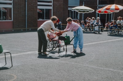 Two people engage in an outdoor activity in a courtyard. One is handing an object to the other in what appears to be a playful setting; a group of children in wheelchairs watch from a distance. The scene is lively with bright sunlight and some colorful parasols providing shade.