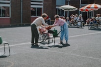 Two people engage in an outdoor activity in a courtyard. One is handing an object to the other in what appears to be a playful setting; a group of children in wheelchairs watch from a distance. The scene is lively with bright sunlight and some colorful parasols providing shade.