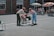 Two people engage in an outdoor activity in a courtyard. One is handing an object to the other in what appears to be a playful setting; a group of children in wheelchairs watch from a distance. The scene is lively with bright sunlight and some colorful parasols providing shade.