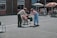 Two people engage in an outdoor activity in a courtyard. One is handing an object to the other in what appears to be a playful setting; a group of children in wheelchairs watch from a distance. The scene is lively with bright sunlight and some colorful parasols providing shade.