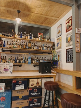 A cozy bar interior with shelves full of various beer bottles displayed. The wall is adorned with vintage beer posters. Boxes of beverages are stacked on the floor beneath the wooden counter. A stool is positioned in front of the bar, and there's ambient lighting from a hanging bulb.