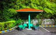 A sheltered seating area in a parking lot featuring a red-roofed structure with four green benches positioned around a central pole. The background showcases dense greenery, including various shrubs and trees, with a concrete wall partially visible. The parking lot is bordered with blue, white, and yellow stripes.
