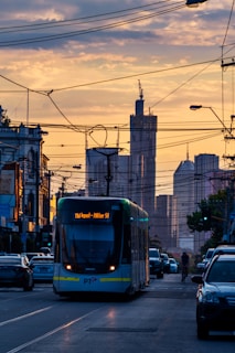 A modern tram is traveling down a city street surrounded by cars, with tall skyscrapers visible in the background under a sky with soft, warm lighting. The roads are intersected by numerous power lines and the overall atmosphere is urban and bustling.