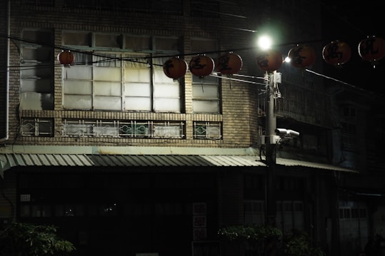 A dimly lit street scene at night featuring a brick building with large windows and a metal awning. Several decorative lanterns with inscriptions hang across the street, illuminated by a streetlight. The setting has a quiet and somewhat eerie atmosphere, enhanced by the shadows and the sparse lighting.