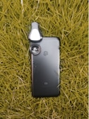 A smartphone lies on a bed of grass, with a detachable lens accessory attached to its camera, enhancing its photographic capabilities.