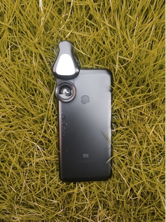 A smartphone lies on a bed of grass, with a detachable lens accessory attached to its camera, enhancing its photographic capabilities.