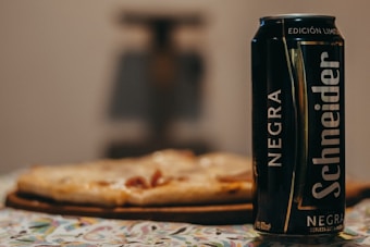 A can of dark beer labeled 'Schneider Negra' is placed on a table. The background features a blurred view of a pizza, suggesting a relaxed dining setting.