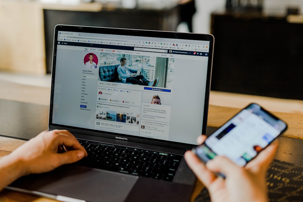 A laptop screen showing a user on Facebook whilst holding a phone in their right hand