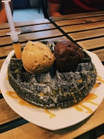 A wooden table holds a plate with a dark waffle topped with two scoops of ice cream, one in a light caramel color and the other in rich chocolate. The waffle is drizzled with a caramel sauce and garnished with powdered sugar. A syringe filled with a matching caramel sauce is embedded in the side, adding an unconventional element to the dessert presentation.