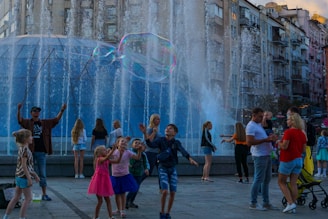 Children and adults are gathered around a fountain, enjoying giant bubbles being made by a performer using long wands. The setting is urban, with tall residential buildings in the background, and people seem engaged and joyful in activities taking place in the plaza.
