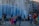 Children and adults are gathered around a fountain, enjoying giant bubbles being made by a performer using long wands. The setting is urban, with tall residential buildings in the background, and people seem engaged and joyful in activities taking place in the plaza.