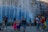 Children and adults are gathered around a fountain, enjoying giant bubbles being made by a performer using long wands. The setting is urban, with tall residential buildings in the background, and people seem engaged and joyful in activities taking place in the plaza.