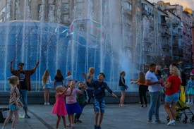 Children and adults are gathered around a fountain, enjoying giant bubbles being made by a performer using long wands. The setting is urban, with tall residential buildings in the background, and people seem engaged and joyful in activities taking place in the plaza.