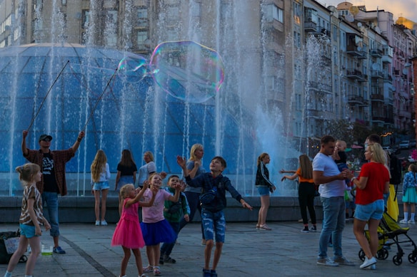 Children and adults are gathered around a fountain, enjoying giant bubbles being made by a performer using long wands. The setting is urban, with tall residential buildings in the background, and people seem engaged and joyful in activities taking place in the plaza.