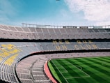 A large, empty football stadium with rows of seats in the stands. The seats form a pattern with the words 'MES QUE UN CLUB' visible. The grass on the field is neatly maintained with alternating light and dark green stripes.