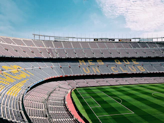A large, empty football stadium with rows of seats in the stands. The seats form a pattern with the words 'MES QUE UN CLUB' visible. The grass on the field is neatly maintained with alternating light and dark green stripes.