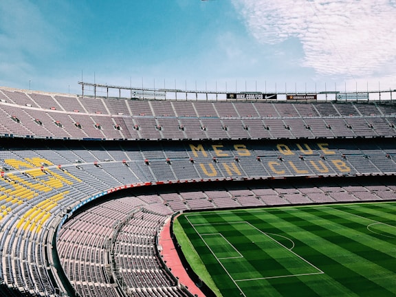 A large, empty football stadium with rows of seats in the stands. The seats form a pattern with the words 'MES QUE UN CLUB' visible. The grass on the field is neatly maintained with alternating light and dark green stripes.