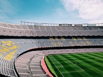 A large, empty football stadium with rows of seats in the stands. The seats form a pattern with the words 'MES QUE UN CLUB' visible. The grass on the field is neatly maintained with alternating light and dark green stripes.