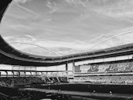 A large sports stadium filled with spectators. The stadium features a curved roof structure and seats filled with people. The sky is partially cloudy, and the field is prepared for an event.