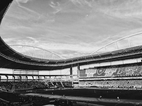 A large sports stadium filled with spectators. The stadium features a curved roof structure and seats filled with people. The sky is partially cloudy, and the field is prepared for an event.