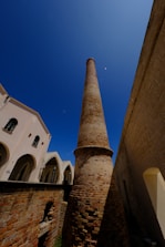 A tall, cylindrical brick chimney against a clear blue sky, surrounded by historic buildings with arched windows and stone walls. The perspective from below emphasizes the height of the chimney.