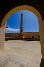 A tall, narrow chimney or smokestack is centrally framed by an arched doorway. The scene includes a courtyard with a stone or concrete floor and walls surrounding the chimney. The sky is clear and bright blue, adding contrast to the structure's earthy tones.