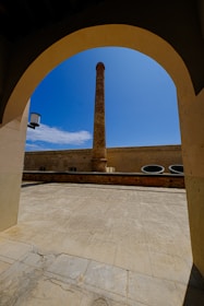 A tall, narrow chimney or smokestack is centrally framed by an arched doorway. The scene includes a courtyard with a stone or concrete floor and walls surrounding the chimney. The sky is clear and bright blue, adding contrast to the structure's earthy tones.