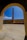 A tall, narrow chimney or smokestack is centrally framed by an arched doorway. The scene includes a courtyard with a stone or concrete floor and walls surrounding the chimney. The sky is clear and bright blue, adding contrast to the structure's earthy tones.