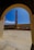 A tall, narrow chimney or smokestack is centrally framed by an arched doorway. The scene includes a courtyard with a stone or concrete floor and walls surrounding the chimney. The sky is clear and bright blue, adding contrast to the structure's earthy tones.