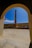 A tall, narrow chimney or smokestack is centrally framed by an arched doorway. The scene includes a courtyard with a stone or concrete floor and walls surrounding the chimney. The sky is clear and bright blue, adding contrast to the structure's earthy tones.